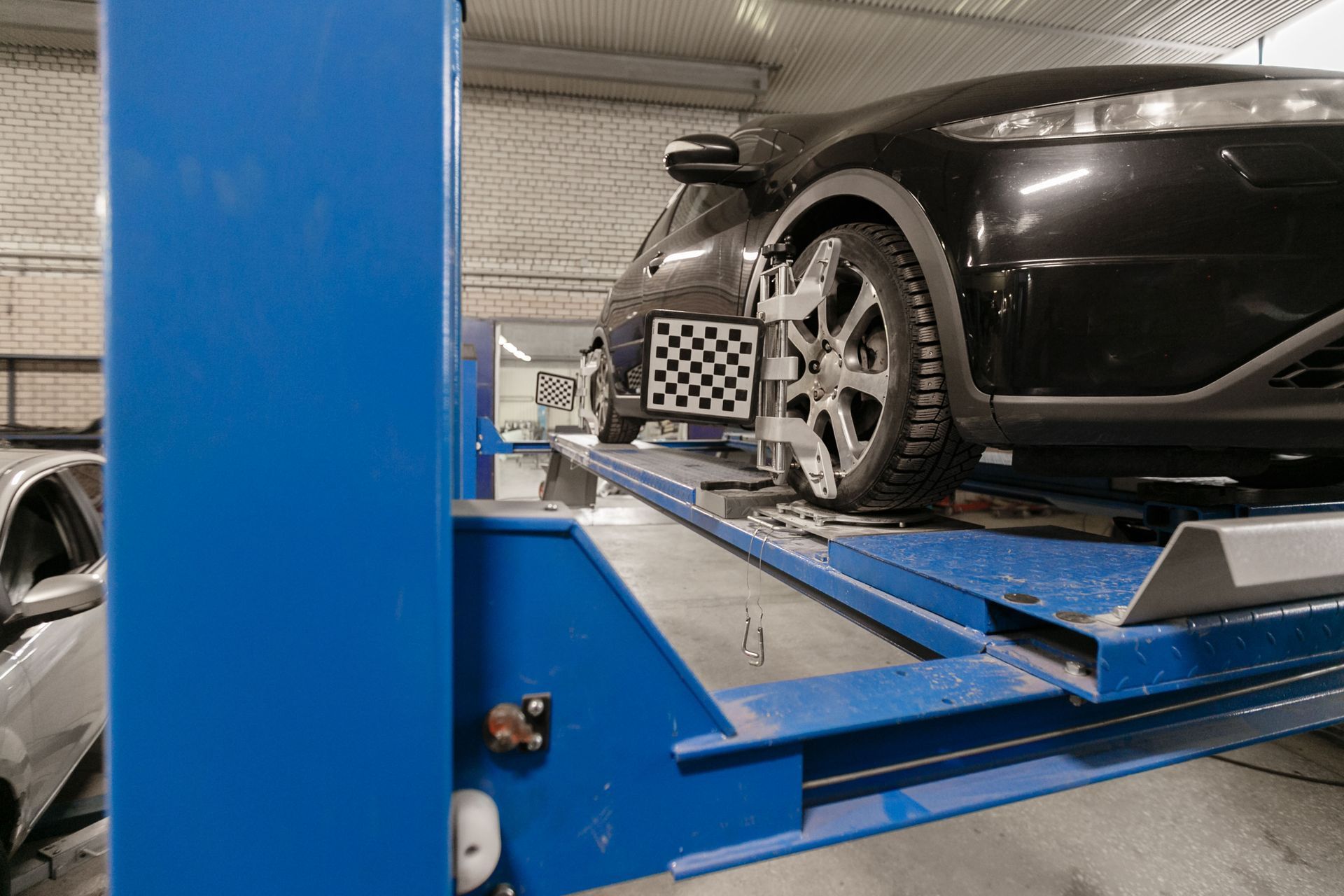 Black car on a lift in a garage, undergoing wheel alignment. The front wheel has a checkerboard target attached.