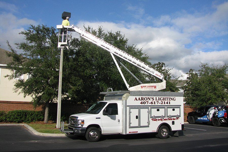 Bucket truck from Adams Lighting raising a worker in a lift beside a tree in a parking lot