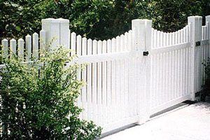 White picket fence with a wavy top and gate beside a leafy green shrub and driveway