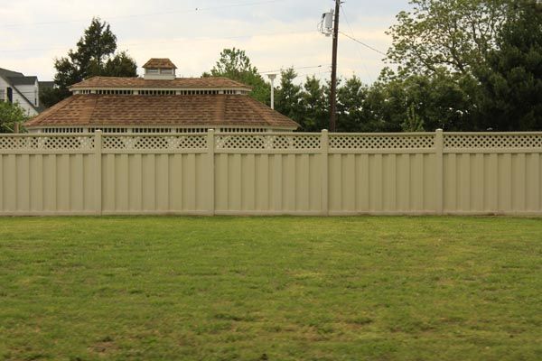 Backyard with green lawn, beige fence, and a brick house roof behind it at dusk