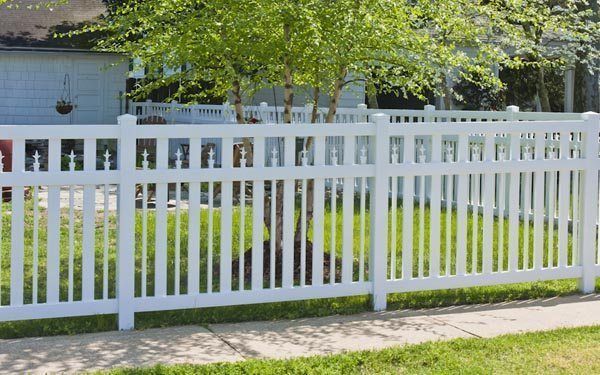 White vinyl picket fence beside a grassy yard with a house and trees in the background.