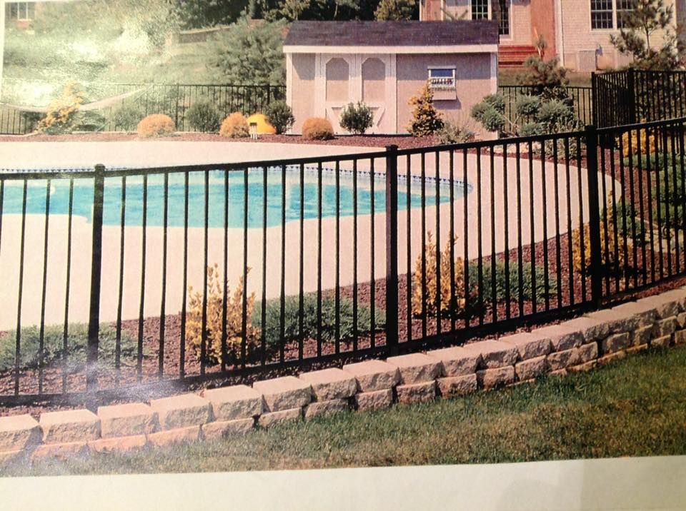Black metal fence around a backyard pool with tan patio and small shed in the background