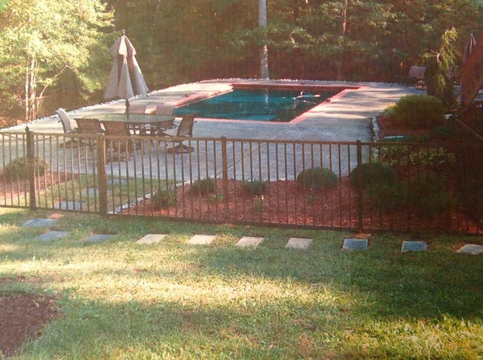 Backyard pool behind black metal fence, with patio chairs and a closed umbrella in sunlight