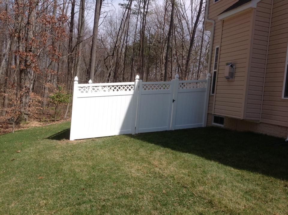 White vinyl fence beside tan house on a grassy yard, with leafless woods in the background