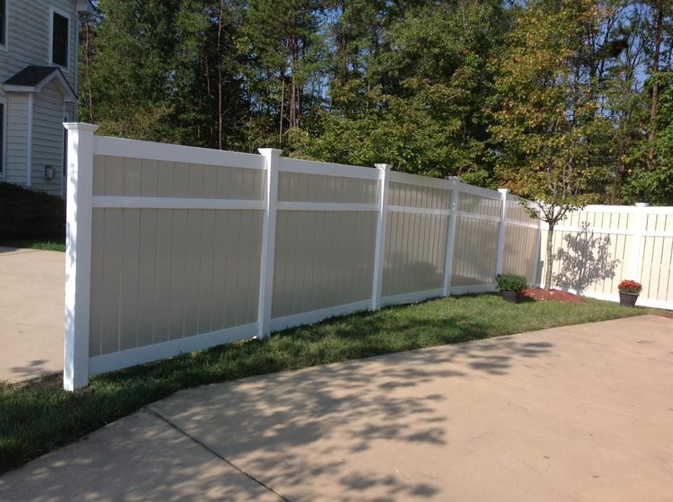 White vinyl privacy fence along a driveway beside a house and lawn, with trees in the background