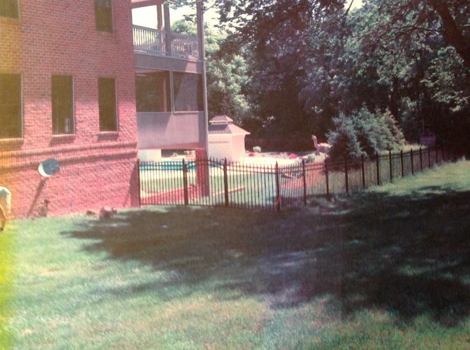 Brick building beside a grassy yard and black fence, with trees casting shade in the background.