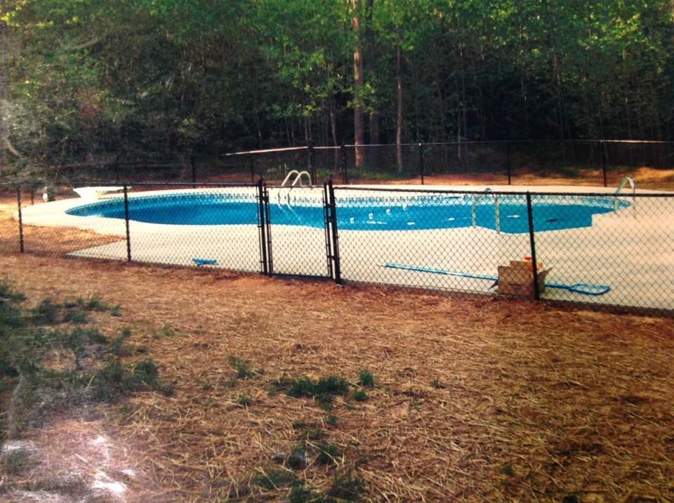 Backyard swimming pool behind a chain-link fence, surrounded by trees and dirt ground.