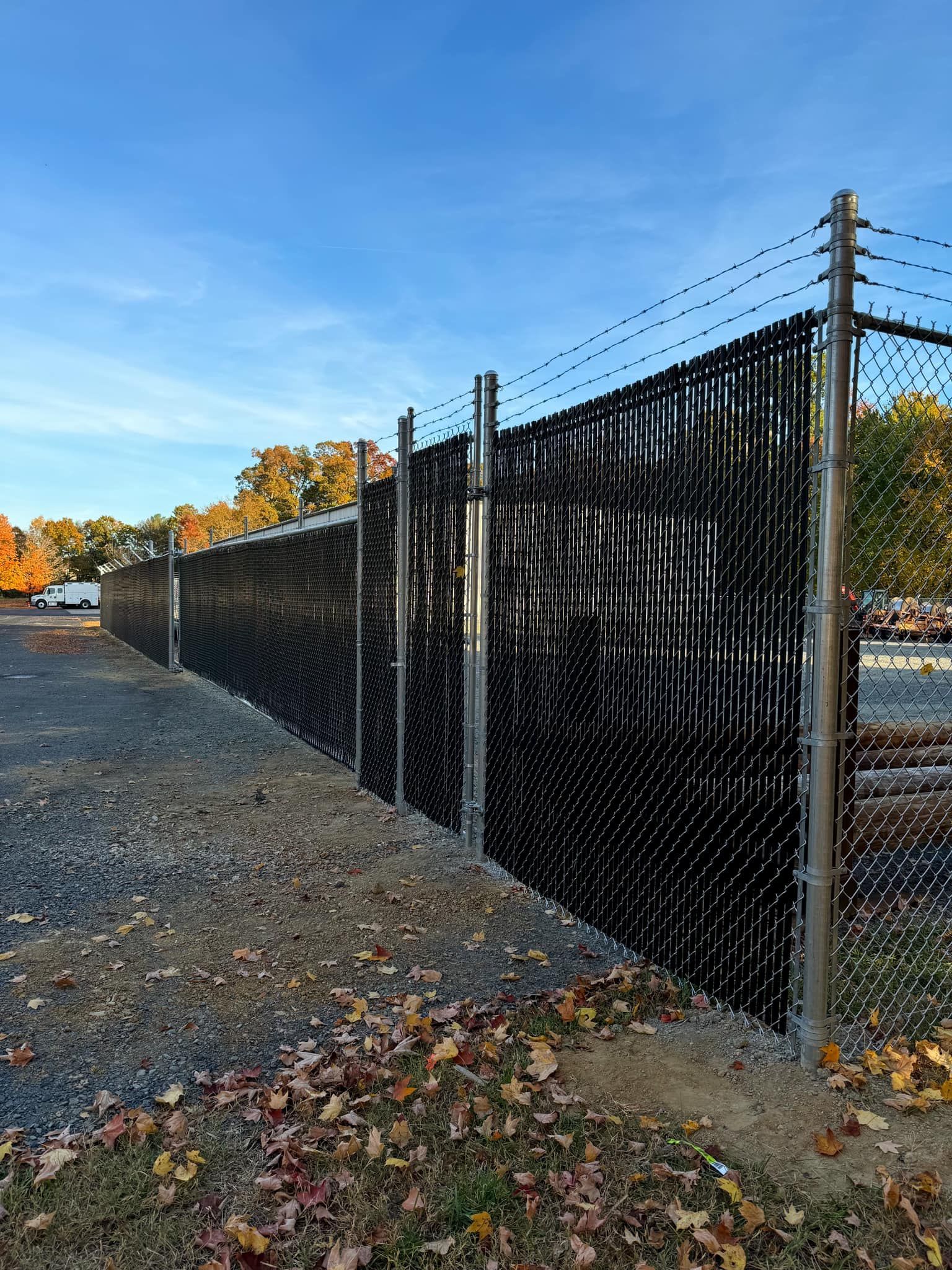 Black chain-link fence with privacy slats along a gravel lot under a blue sky with autumn trees in the background