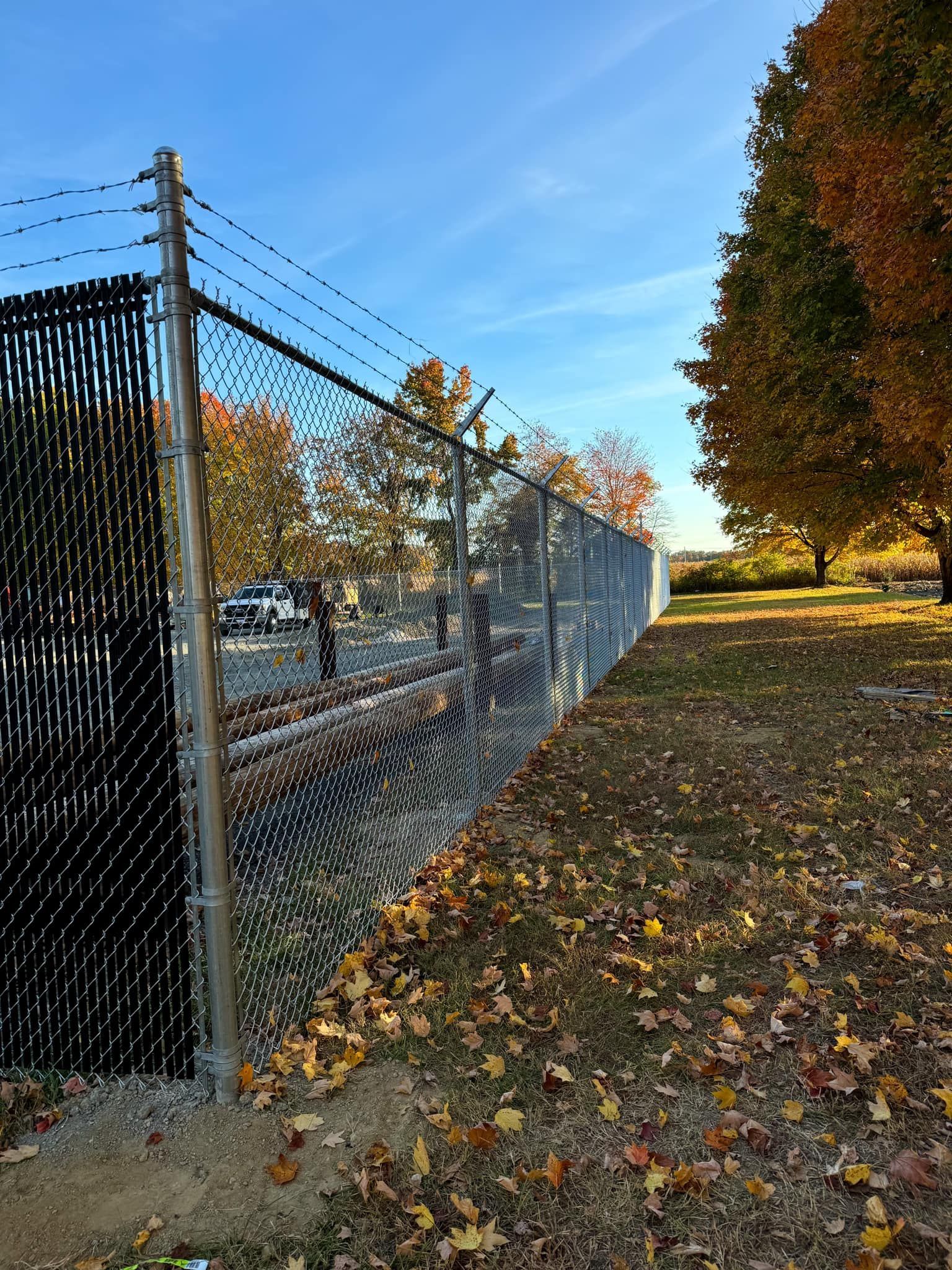 Chain-link fence beside a leaf-covered grassy path in autumn, with trees and blue sky.