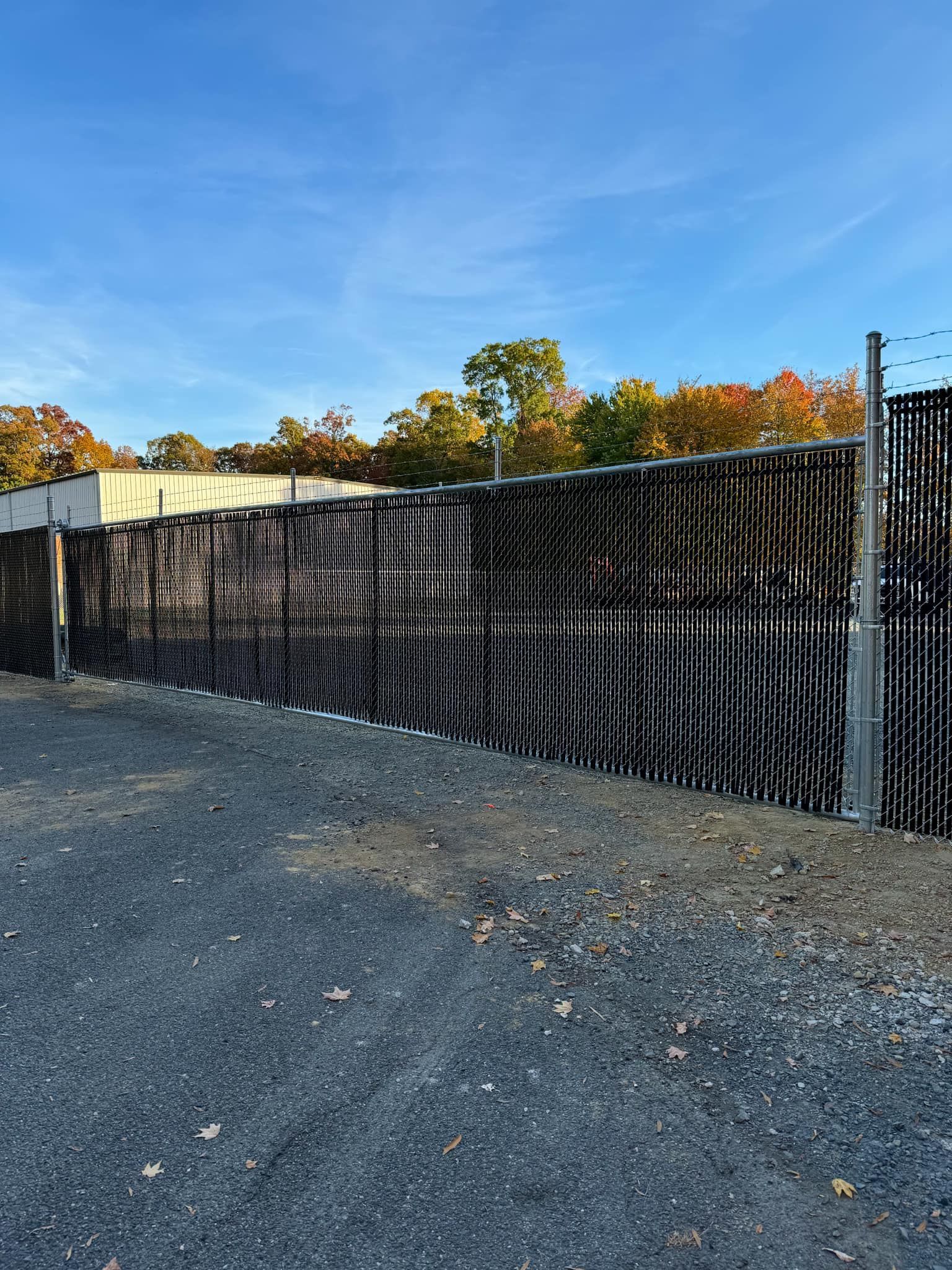 Black mesh fence beside a gravel lot under a blue sky with autumn trees in the background