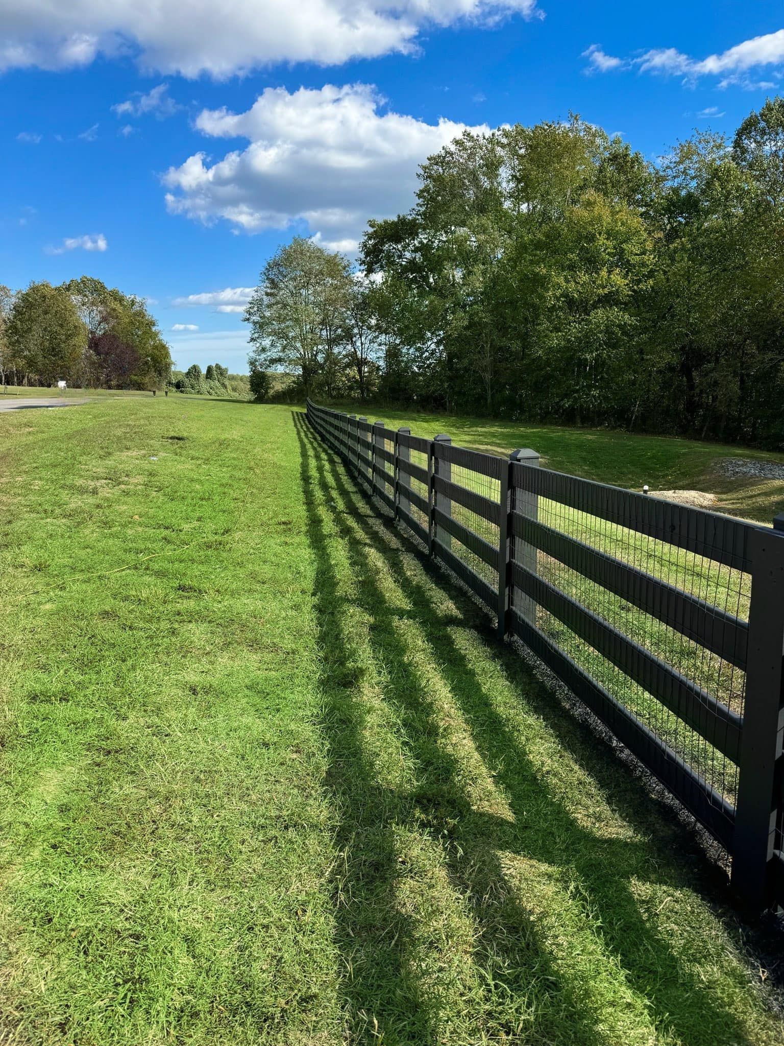 Black wooden fence casting long shadows beside a green field under a blue sky with clouds.