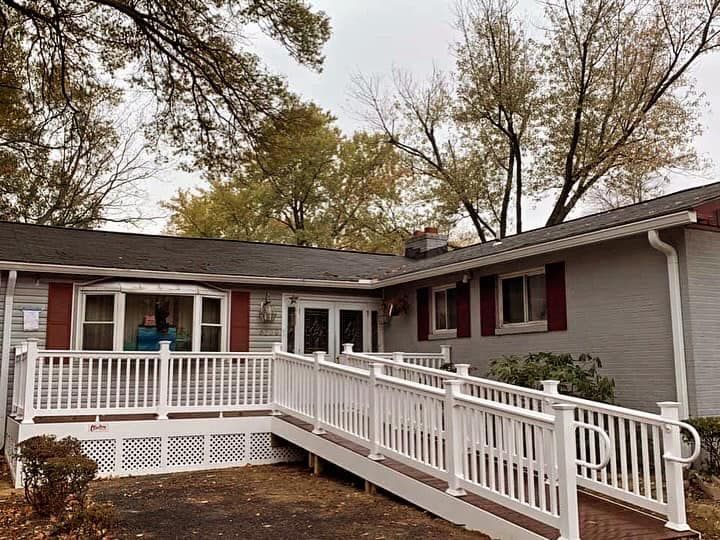 Front porch ramp leading to a beige house with red shutters and white railings beneath autumn trees