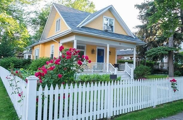 Yellow house with white picket fence and blooming pink flowers by a tree-lined yard