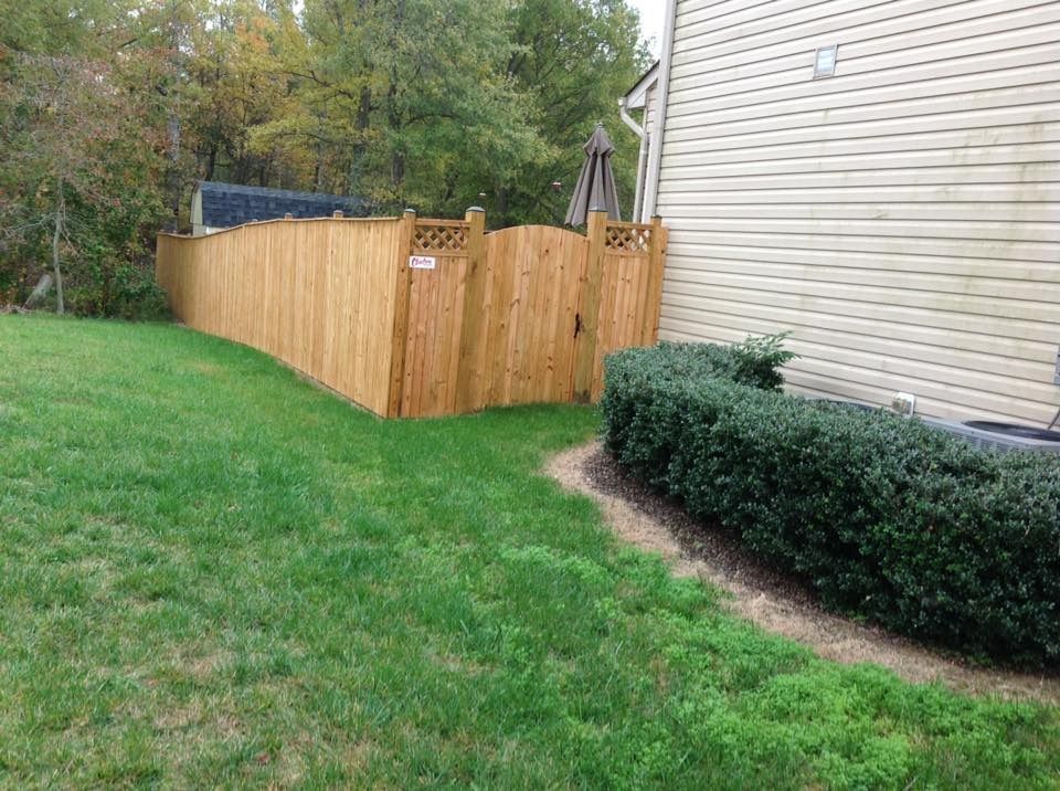 Wooden fenced patio beside a beige house, bordered by green lawn and trimmed shrubs.
