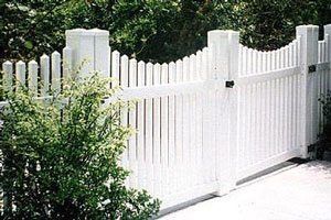 White picket gate and fence beside a driveway, with green bushes and trees behind it.