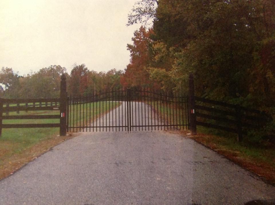 Closed black metal gate blocking a gravel driveway, with trees and grassy fields on both sides