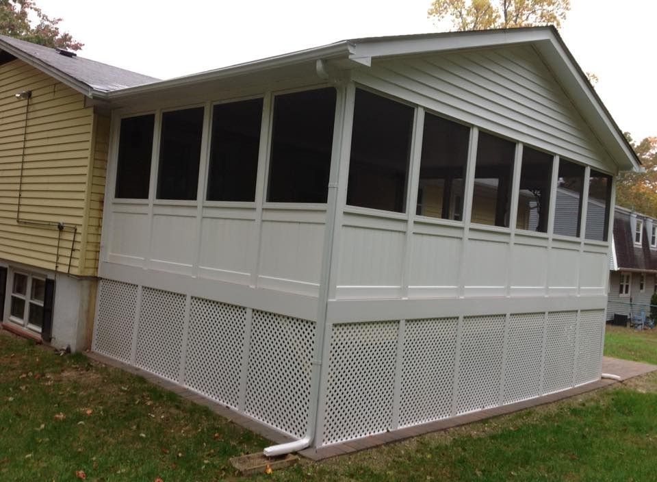 White screened porch or sunroom attached to a beige house, with lattice skirting and dark window panels.
