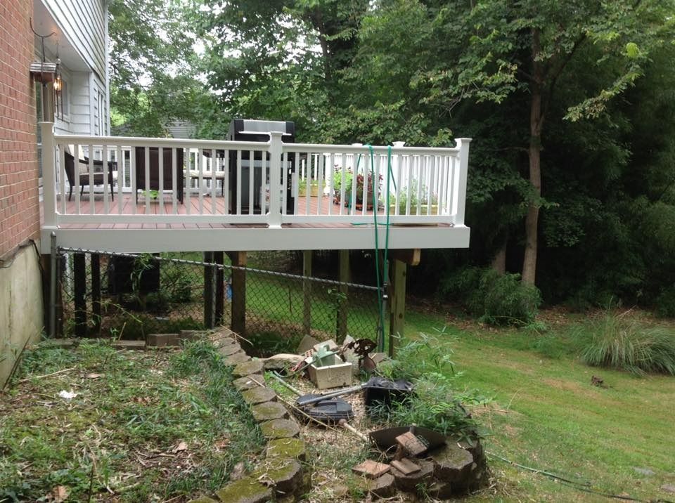 Raised deck with white railing over a sloped backyard, supported by posts and surrounded by trees.