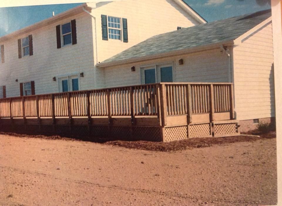 Two-story beige house with a long wooden deck and railing on a gravel lot under a blue sky