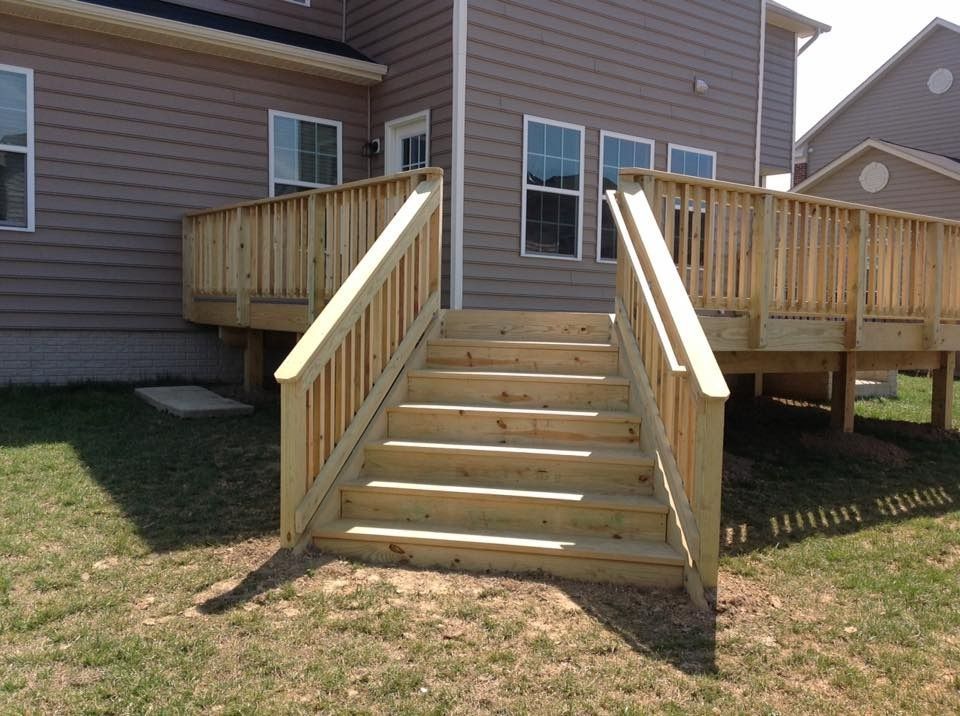 Wooden staircase with railings leading to a raised deck on a house, with a grassy yard below.