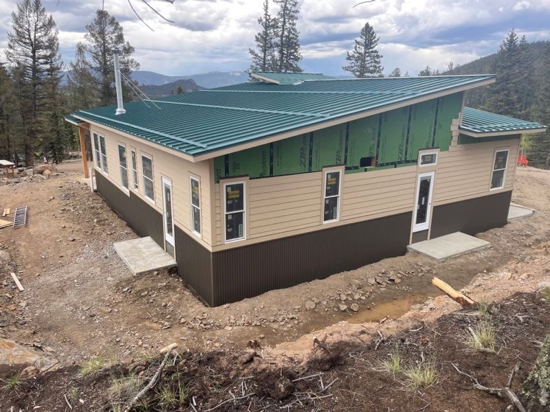 Small beige house with green metal roof on a dirt lot in a wooded mountain area