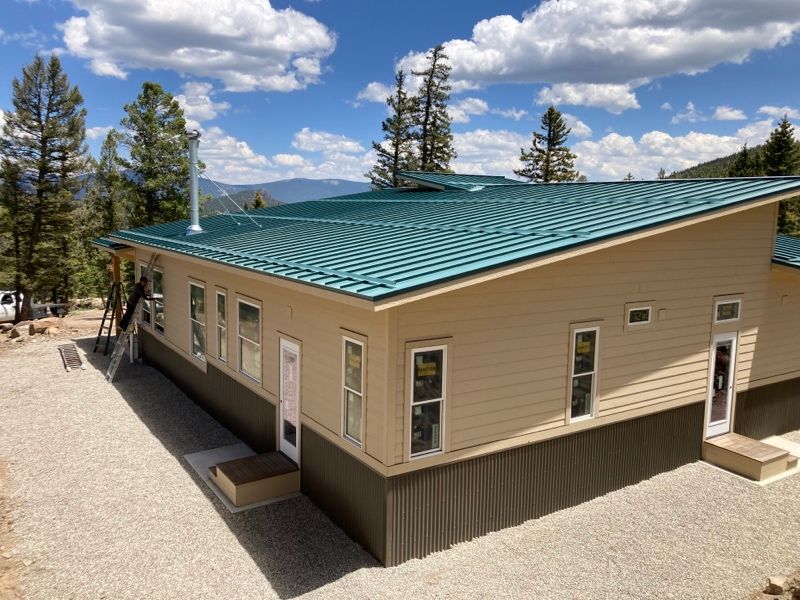 Tan house with teal metal roof in a pine forest under a blue sky with clouds