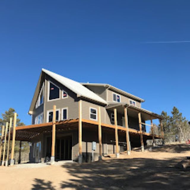 Large house under construction with wraparound porch and wood framing on a sunny, sandy lot