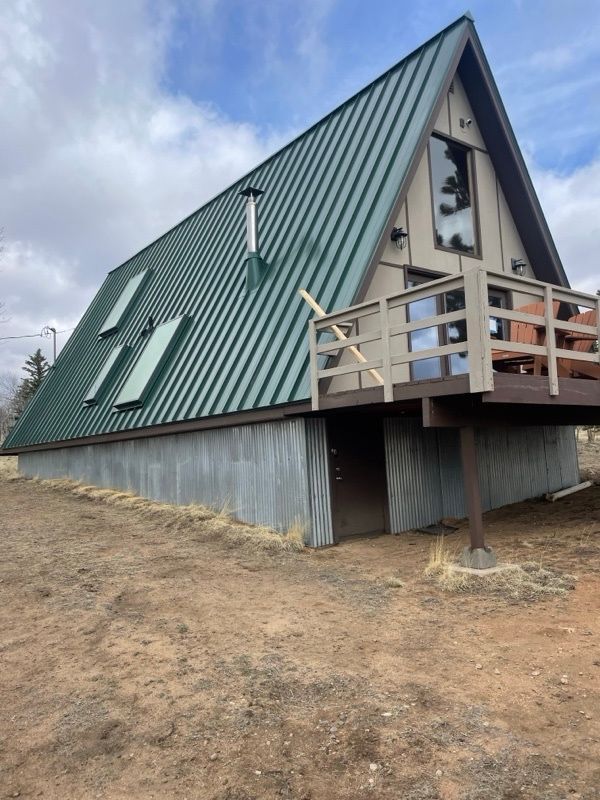 A small A-frame cabin with a green metal roof and wooden balcony on a dirt slope under a cloudy sky.