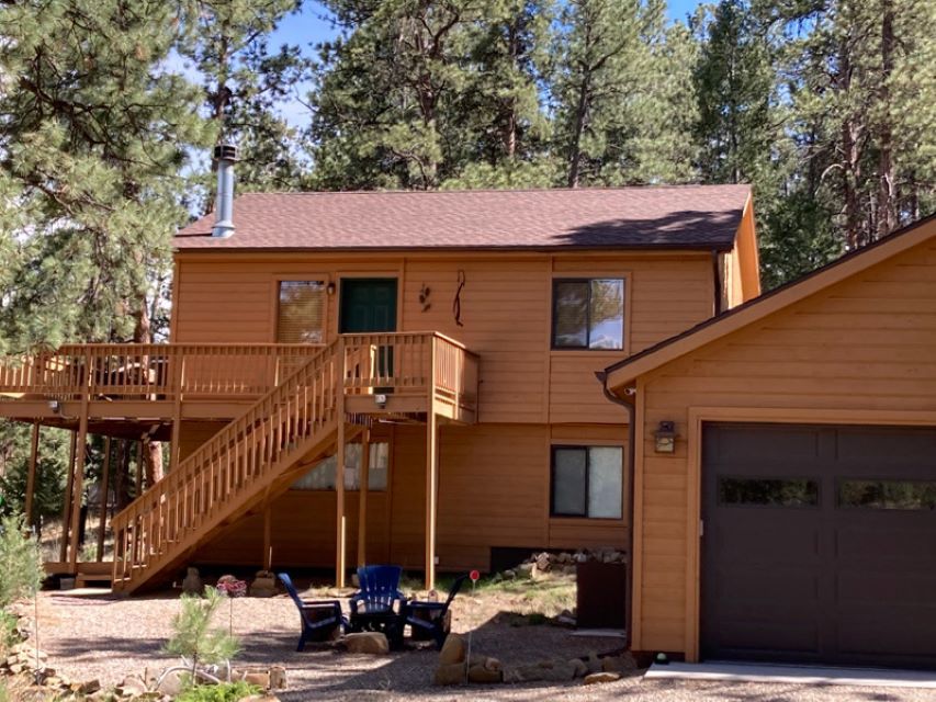 Two-story tan cabin with wooden deck and stairs beside a garage in a pine forest