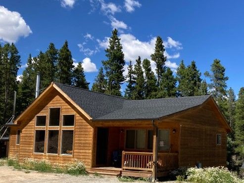 Wooden cabin with a porch and large windows under a blue sky, surrounded by tall pine trees