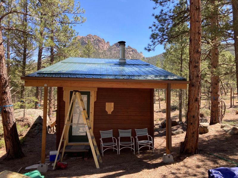 Small wooden cabin in a pine forest with a blue roof and chairs on the porch