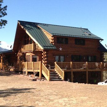 Wooden cabin with green metal roof and wraparound porch on a dry, open lot
