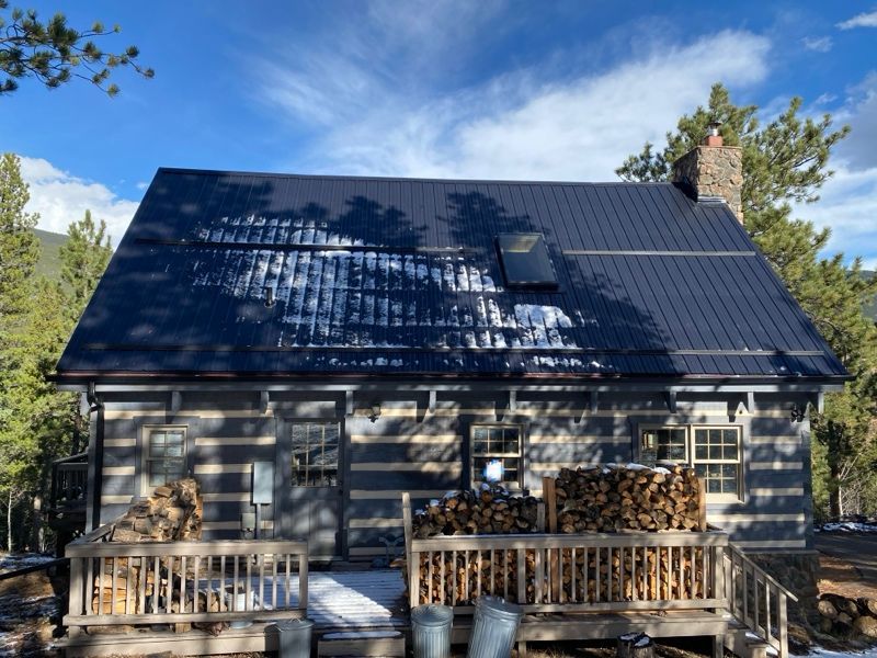 Cabin with dark metal roof, stone chimney, and wooden deck in a wooded mountain setting