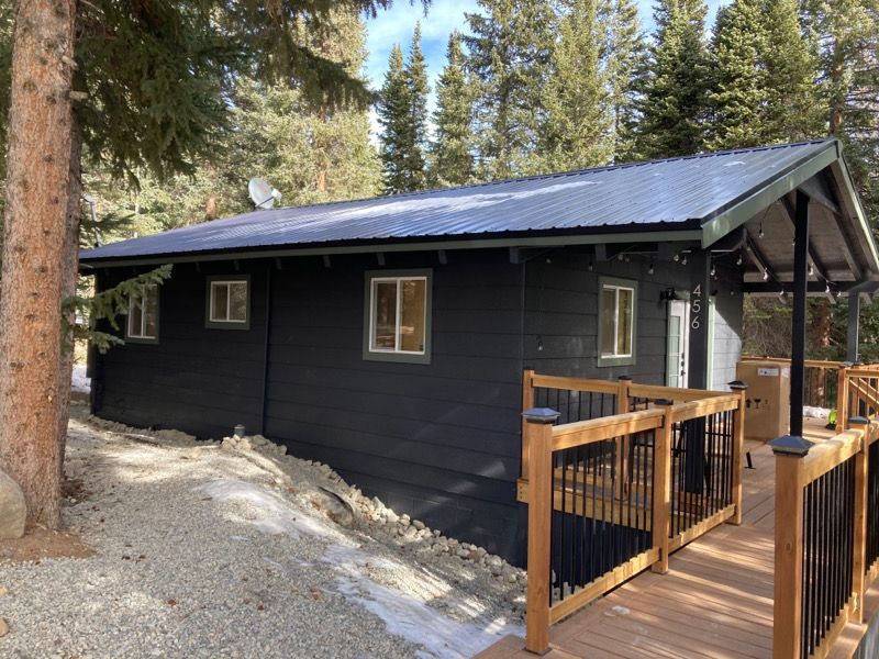 Black cabin with a metal roof and wooden porch railing in a forest clearing