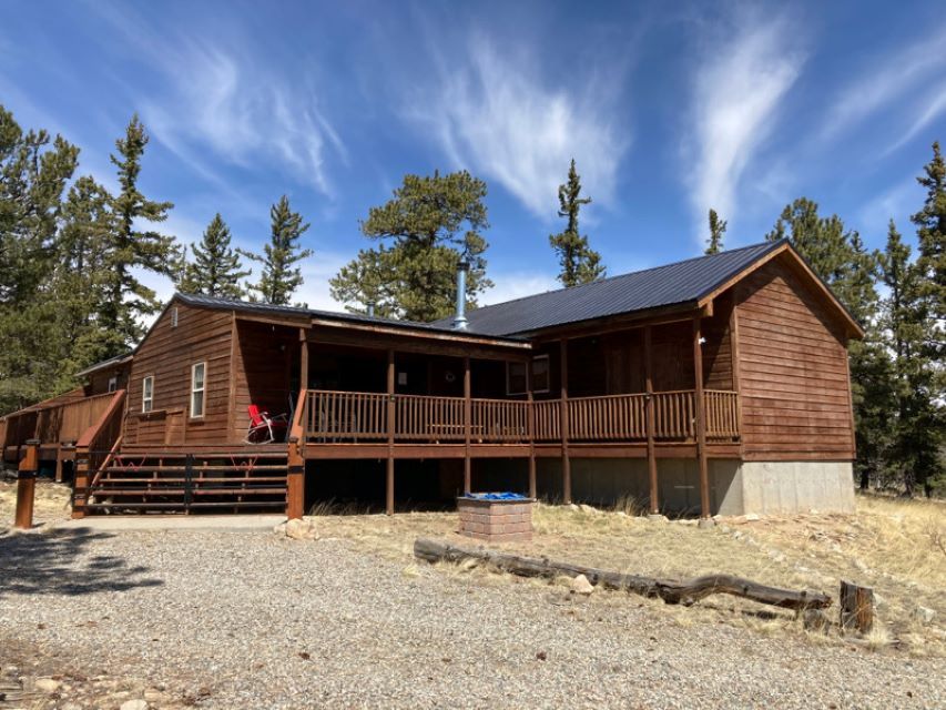 Wooden cabin with a wraparound porch in a pine forest under a blue sky