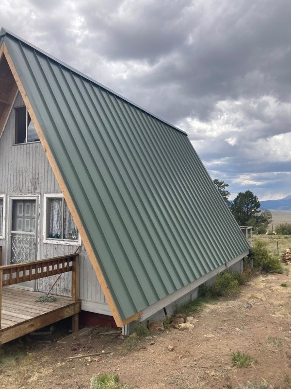 A-frame cabin with a steep green metal roof and wooden deck under cloudy skies.