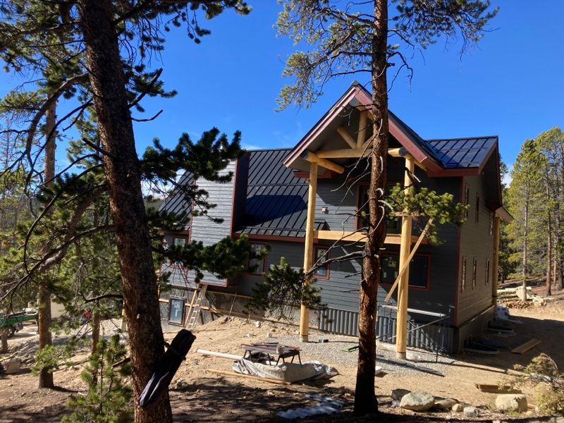 Wooden cabin under construction on a wooded hillside, with a dark roof and pine trees in front.