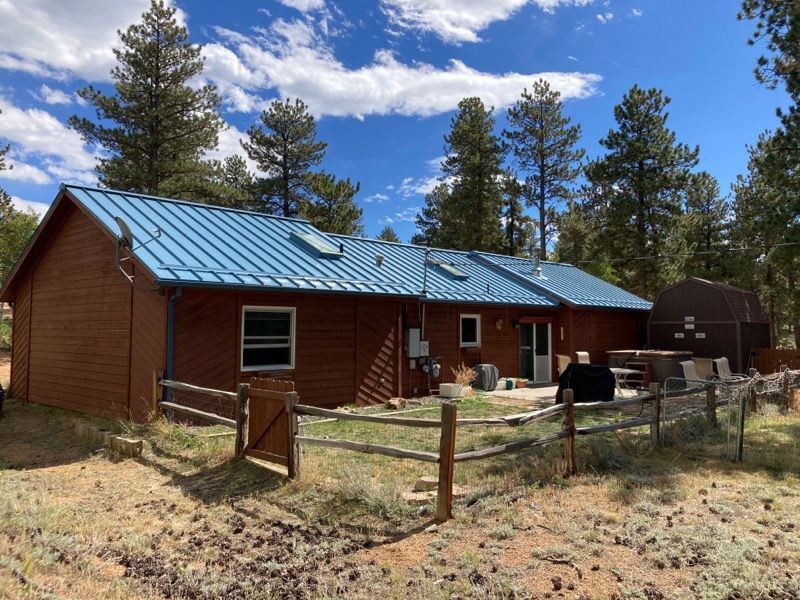 Brown cabin with blue metal roof behind a wooden fence in a pine forest under a partly cloudy sky