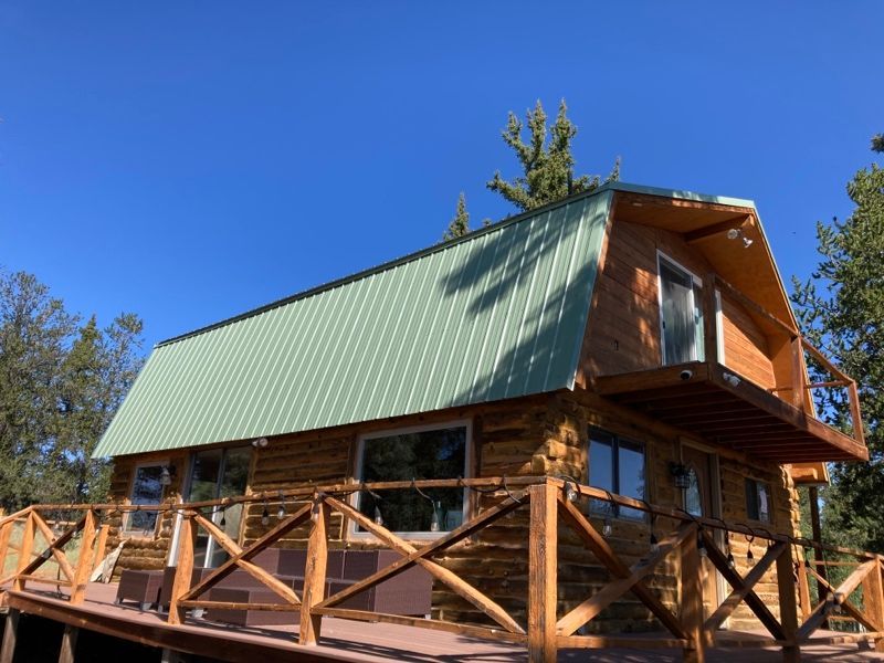 Wooden cabin with green metal roof and wraparound porch amid tall trees under a clear blue sky