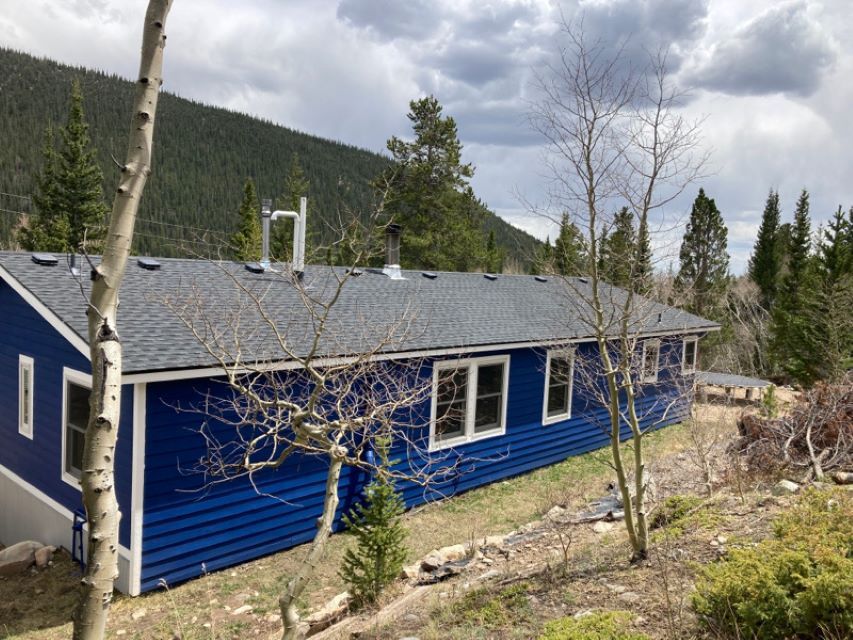 Blue cabin with gray roof in a wooded mountain area under cloudy sky