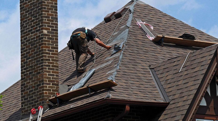 Worker repairing a shingled roof beside a brick chimney, with tools and exposed underlayment visible