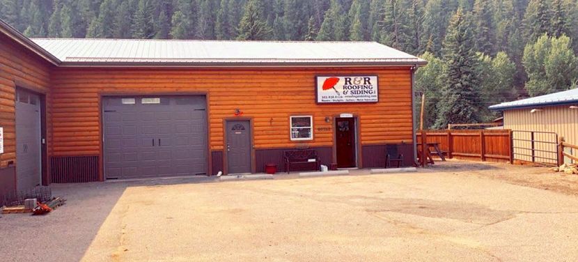 Small red-brown industrial building with a garage door, sign, and fenced gravel lot in a wooded area
