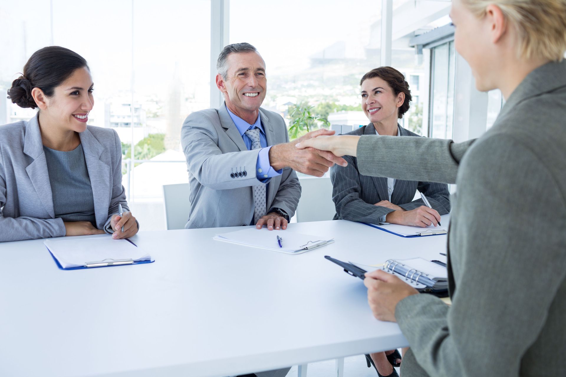 Business people shaking hands at a table, smiling, in a bright office.