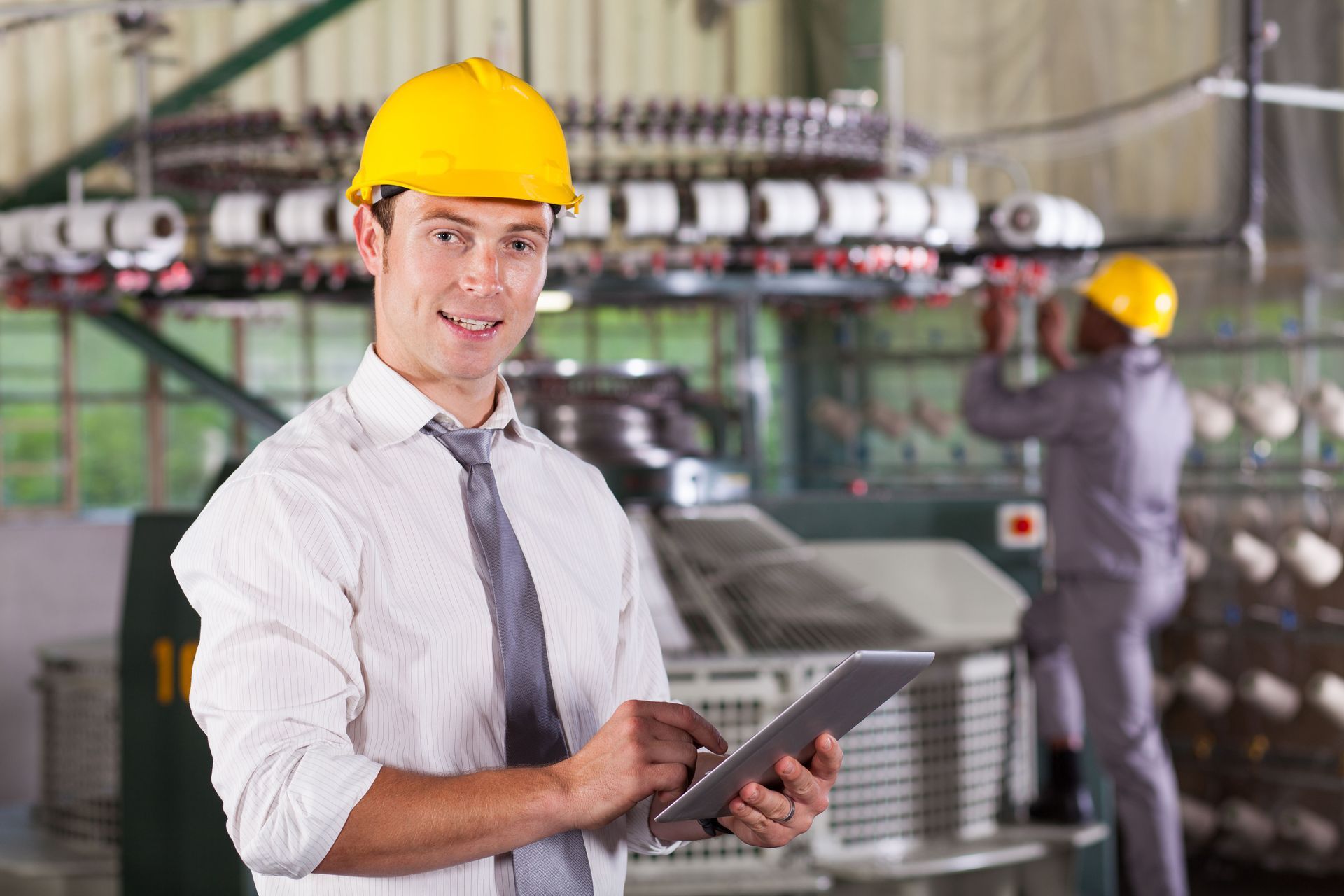 Man in a hard hat using a tablet in a textile factory, another worker in background.