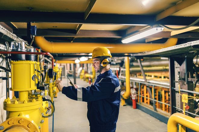 Worker in yellow hard hat and hearing protection checks machinery in a yellow-piped industrial setting.