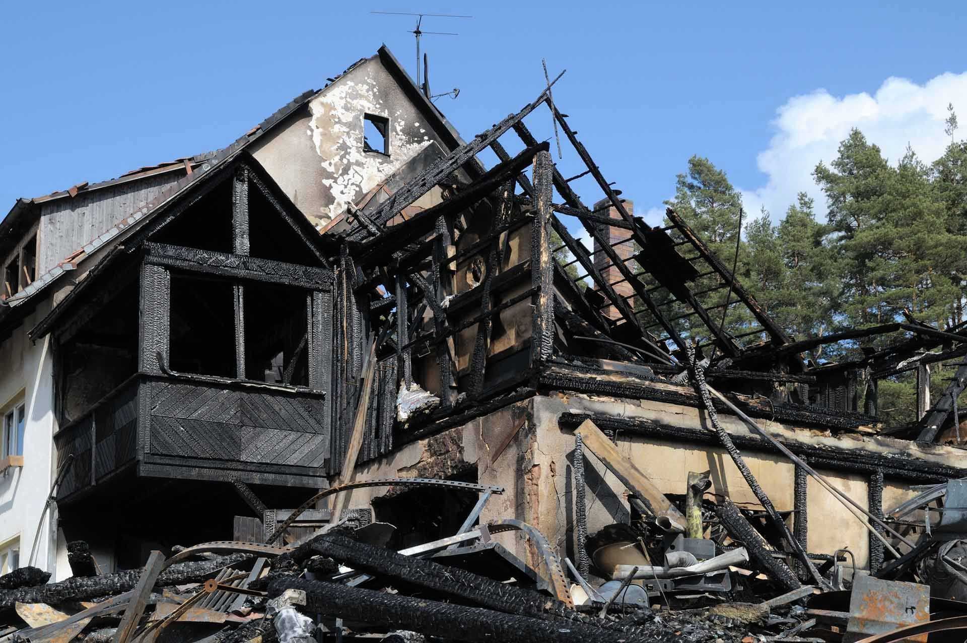 A house destroyed by fire; charred wood and rubble visible; blue sky background.