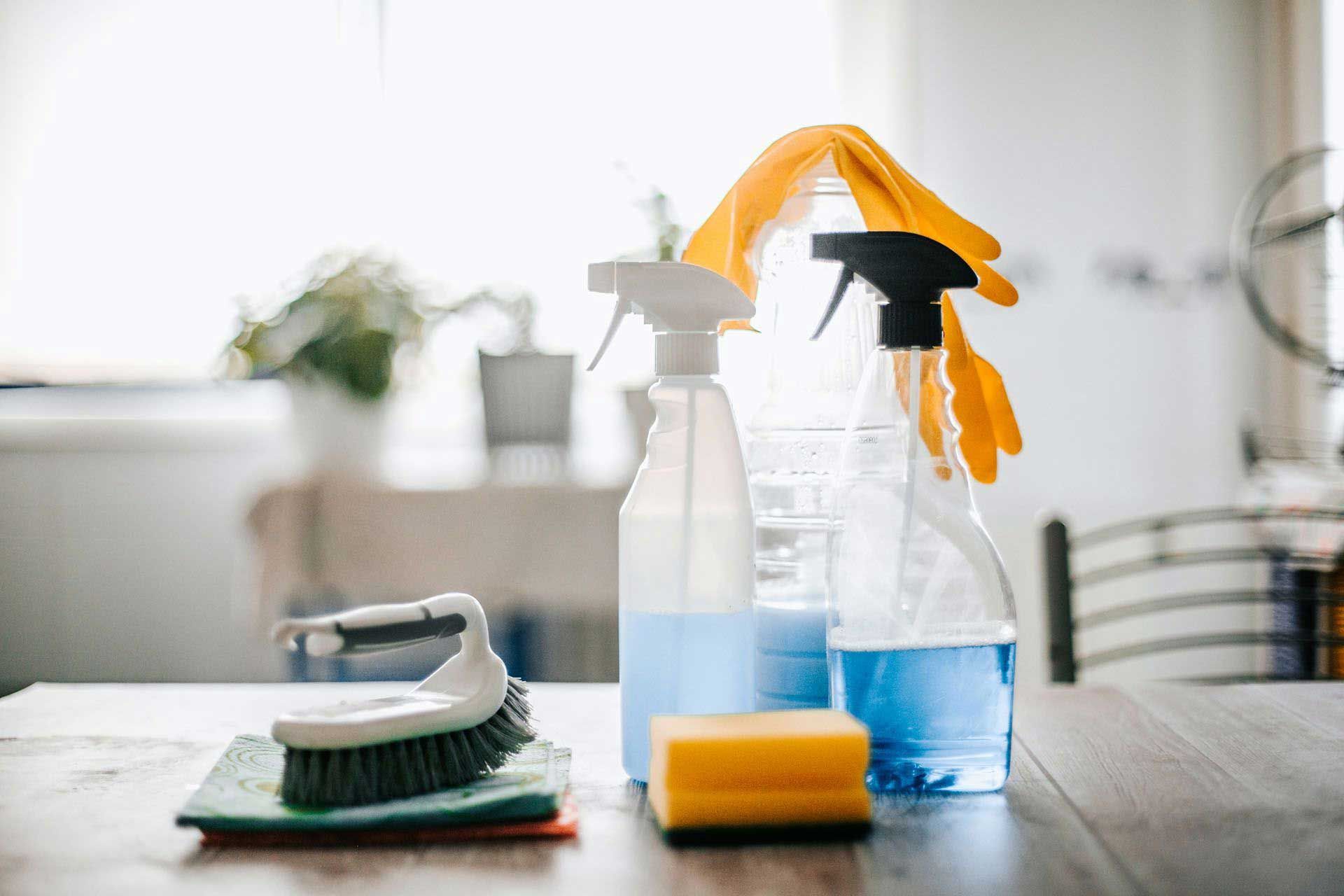 Cleaning supplies on a wooden table: spray bottles, gloves, scrub brush, and sponge.