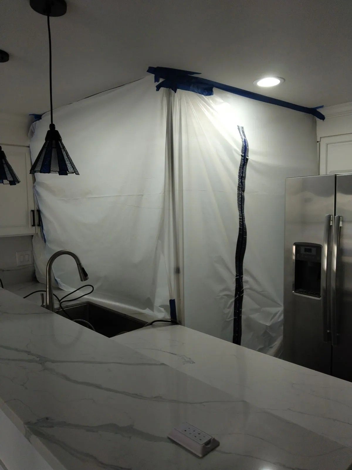 Kitchen with white containment barrier during construction. Counter, sink, and refrigerator visible.