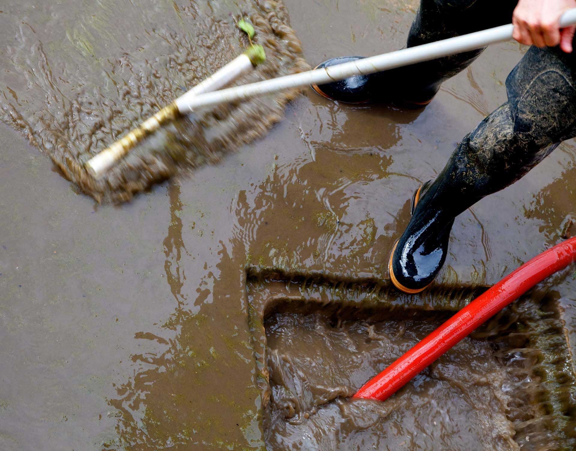 Person in black boots clearing debris with a rake from a flooded street drain.