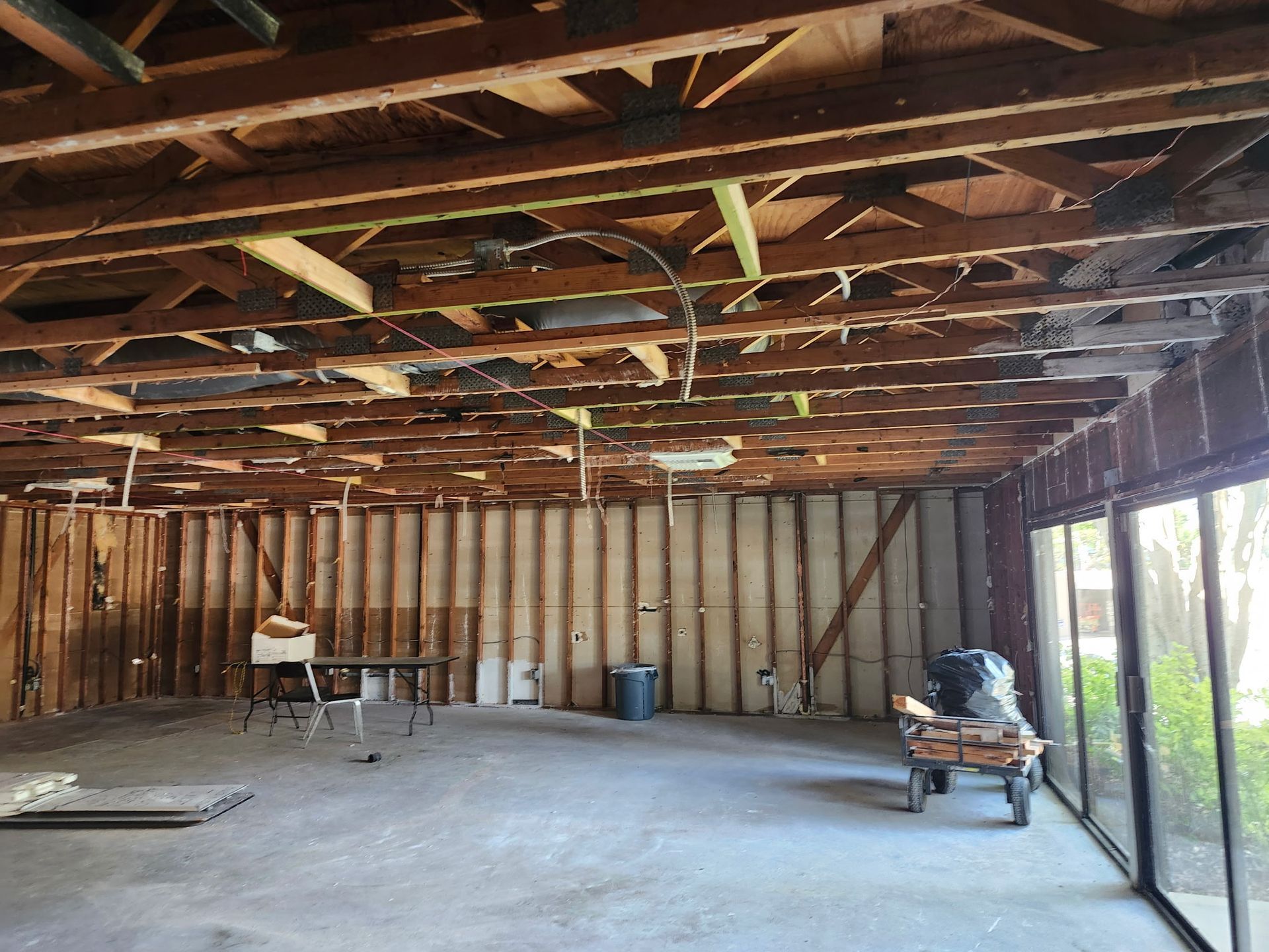 Interior of a gutted building with exposed ceiling beams and wall studs. A concrete floor and large windows are visible.